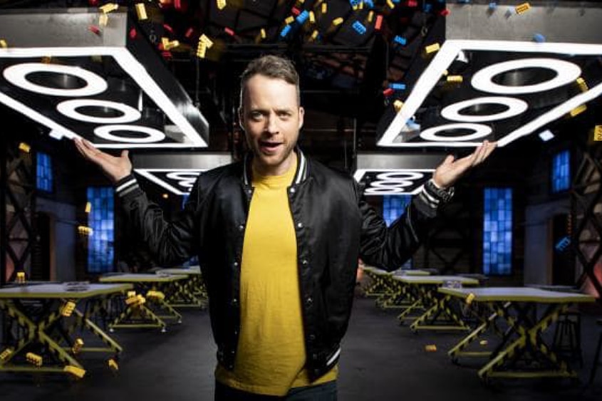 A man is standing in front of a series of tables with a large Lego piece in the background 